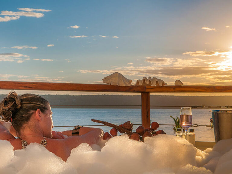 A woman with sparkling wine having a bubble bath outside while overlooking the ocean
